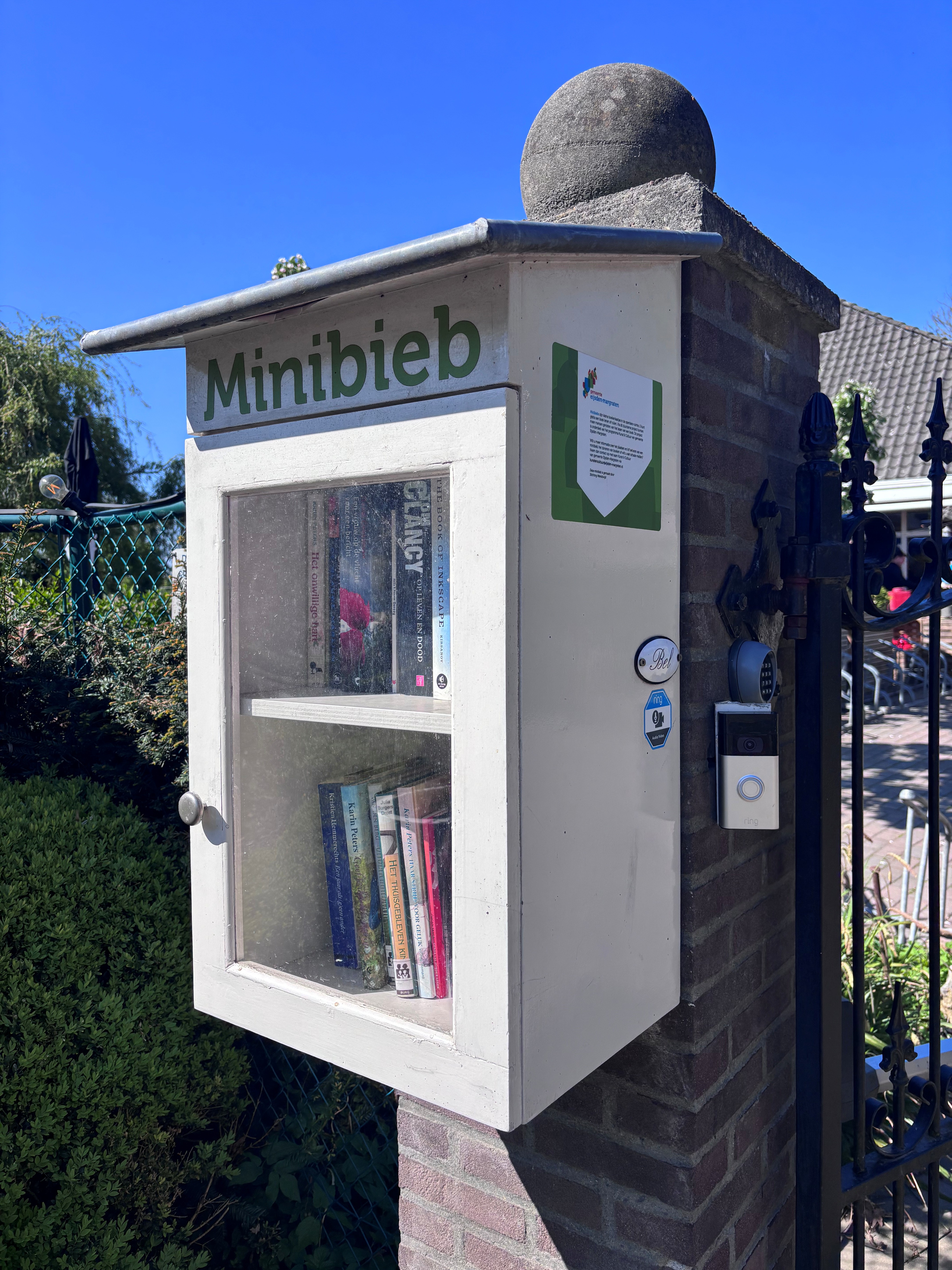 witte minibieb met boeken erin hangt aan een muurtje buiten langs een woning
