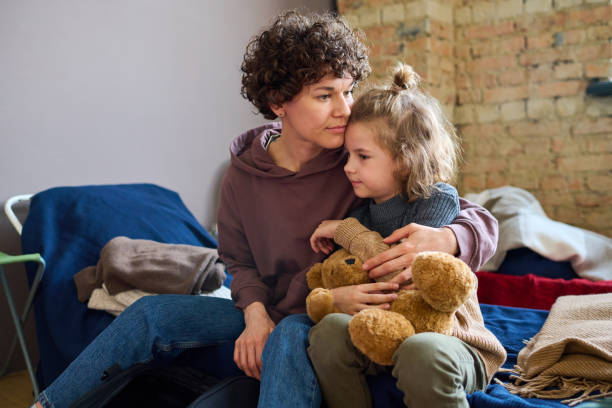 Een moeder en dochter zitten naast elkaar op bed waarbij moeder de dochter vast heeft en dochter een knuffelbeer. 