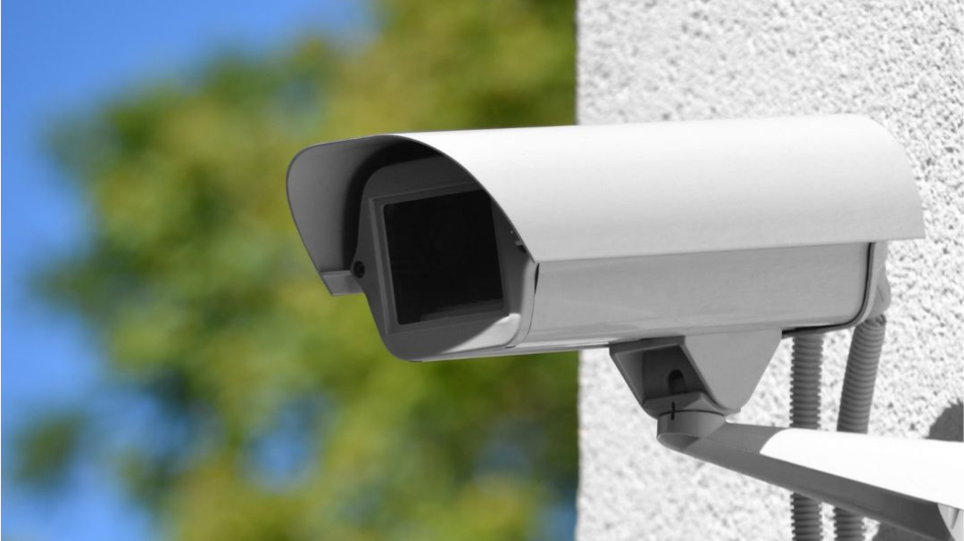 Camera op witte buitenmuur en bomen op achtergrond. 